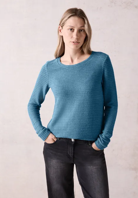 Frau mit langen blonden Haaren trägt blauen Pullover und schwarze Jeans, steht vor hellem Hintergrund.